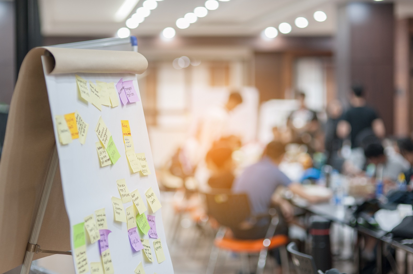 Flipchart mit bunten Notizzetteln vor sitzenden Personen am Tisch