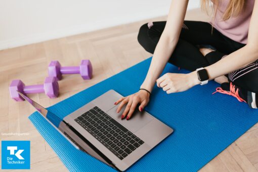 Person sitzt in Sportsachen auf einer Sportmatte und schaut auf den Laptop.