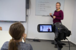 Eine Person erklärt etwas vor einem beschriebenen Whiteboard und wird dabei gefilmt.