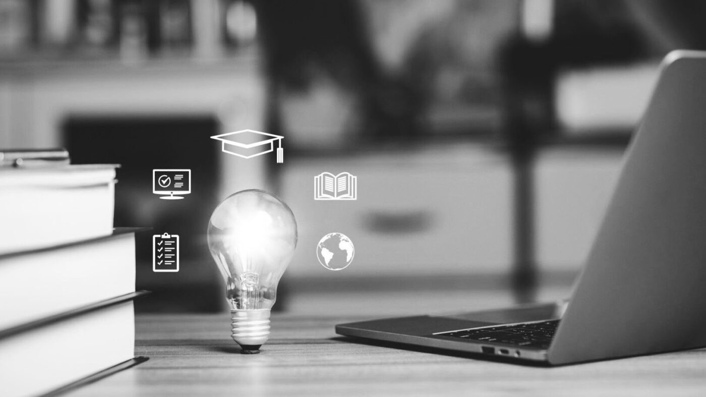 Schreibtisch mit Laptop und Büchern. Im Vordergrund eine Glühbirne, die Icons für digitale Bildung umgibt.